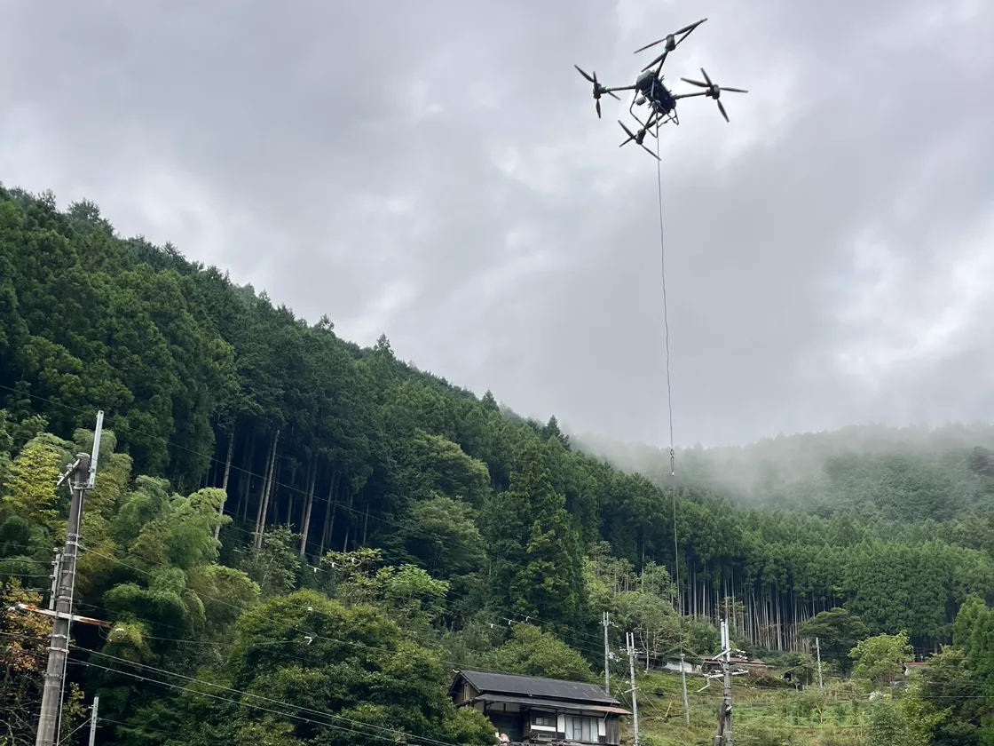 山あいの集落の上空で、ロープの先にワイヤーを吊り下げて飛行するDJI FlyCart 30