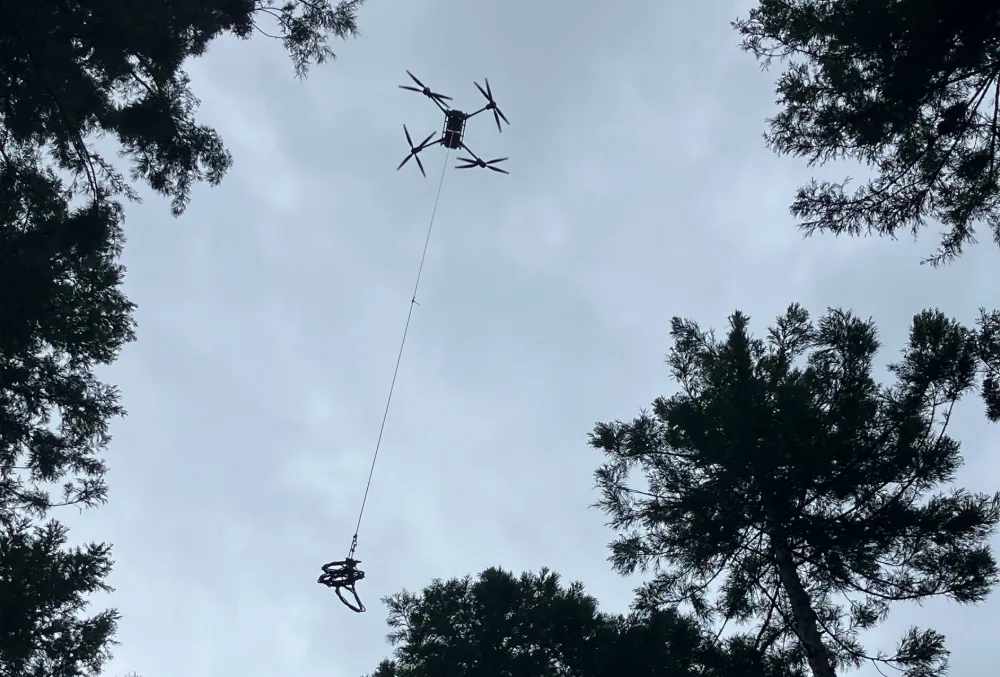 高い木々の間から見上げた、ワイヤーを吊り下げて飛行するFlyCart 30